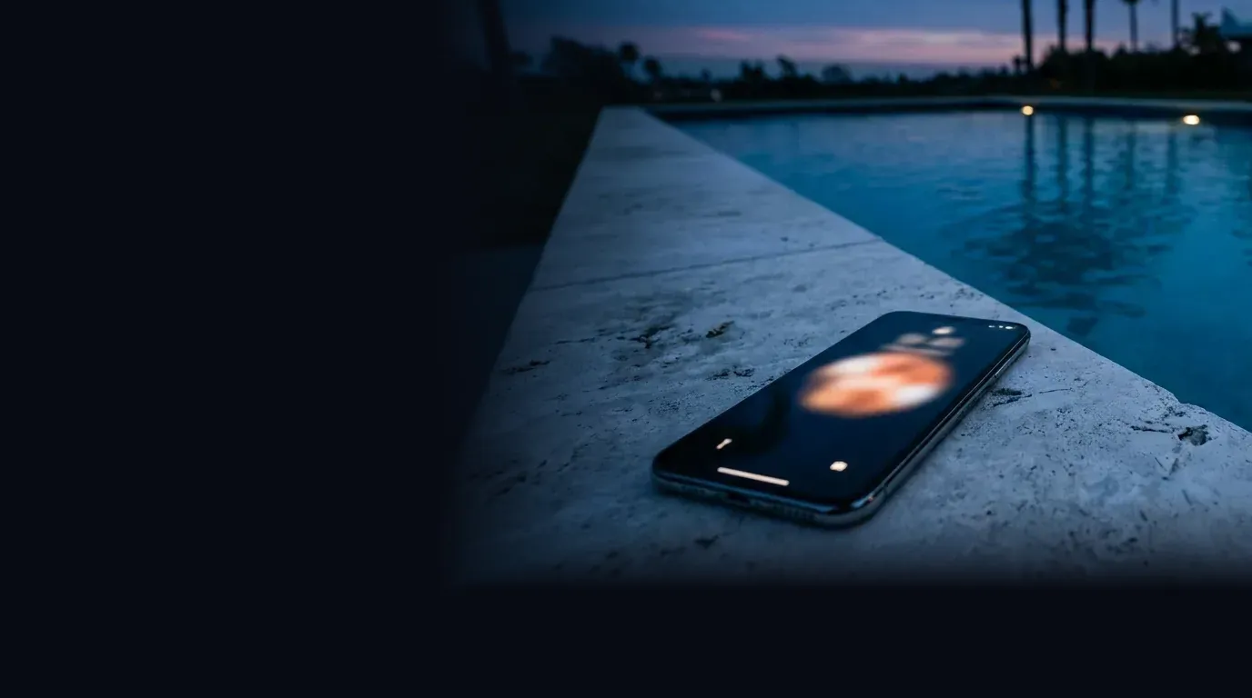 A smartphone with a glowing incoming call screen lying on the stone coping of a lit custom pool at dusk