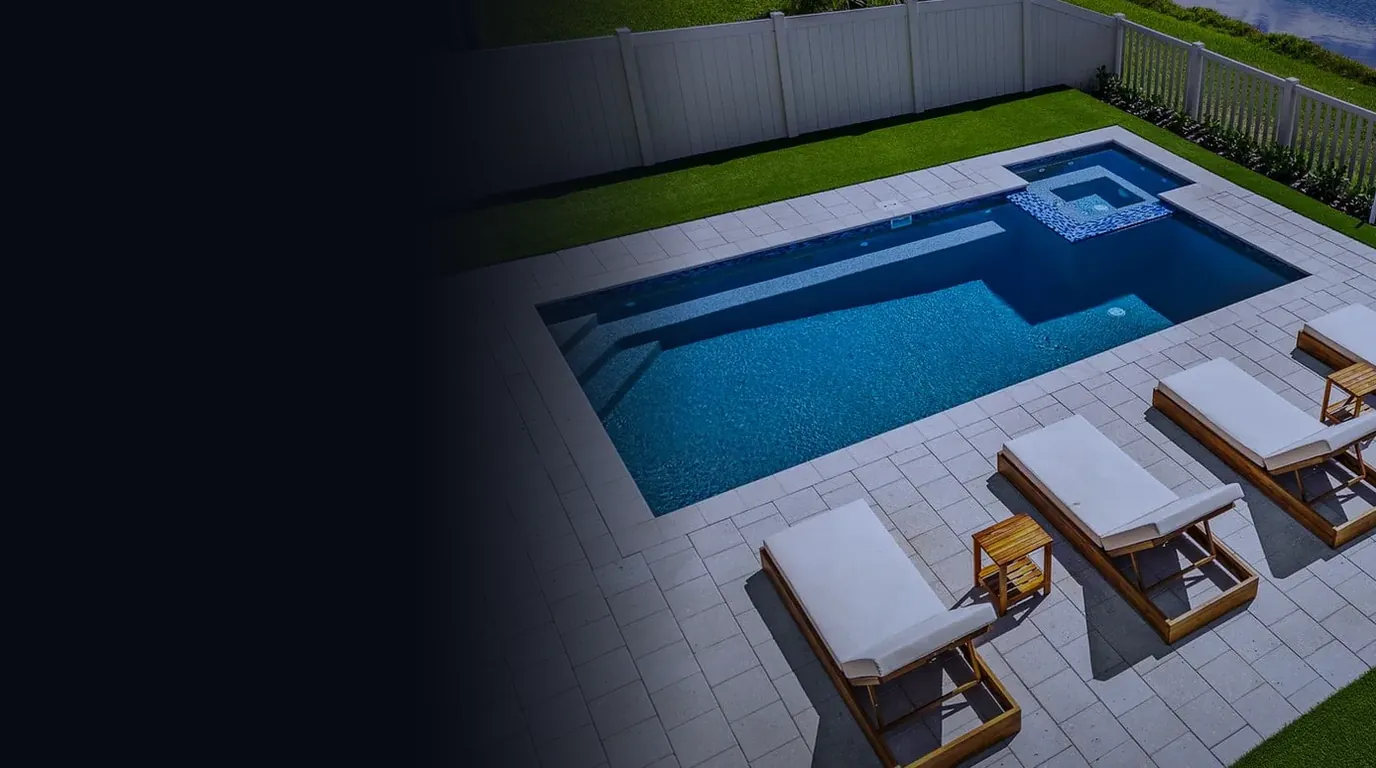 Aerial view of a finished rectangular pool with an attached spa, a paver patio and four lounge chairs, with a fence and a lake behind