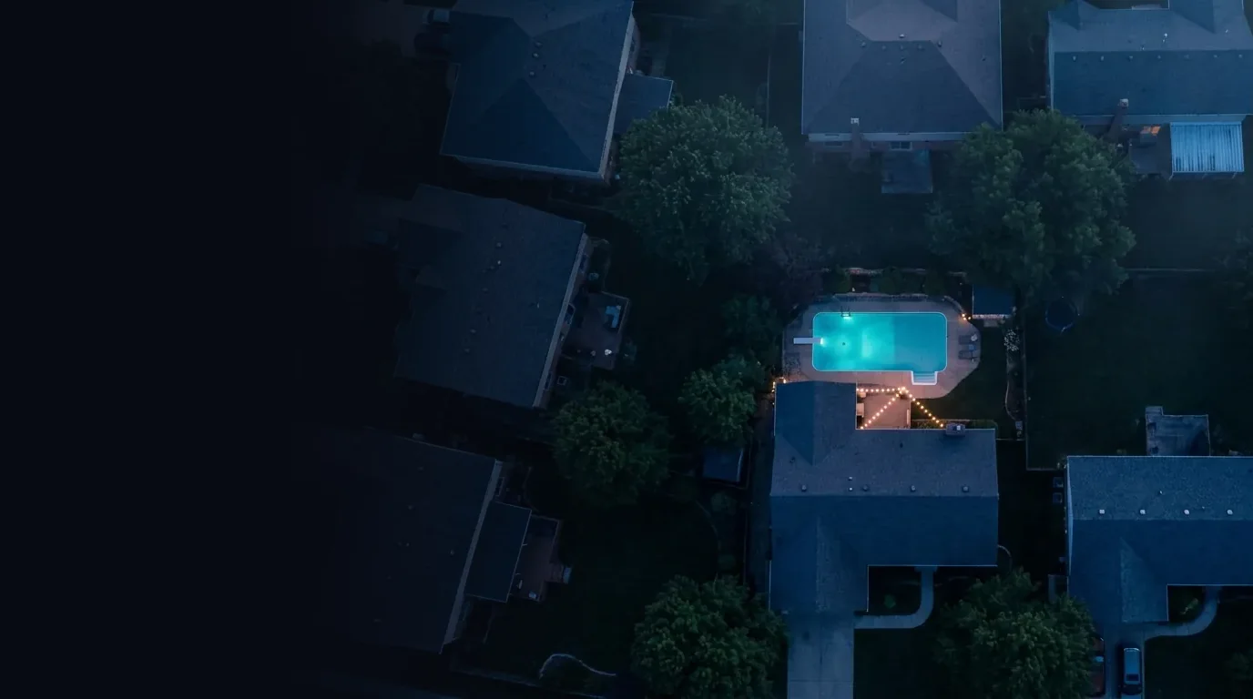 Drone view of a neighborhood at dusk with one lit backyard pool standing out among the rooftops
